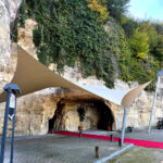 Beige and white tensile canopy over a red carpet leading to a rocky cave entrance beneath an ivy-covered cliff.