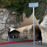 Beige tensile canopy over a cave entrance in a rocky wall, with a red carpet along the pavement and a sign reading 'CAVERNE' nearby.