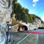 Rocky cliff with green vegetation and a shaded cave entrance under beige fabric canopies; a red carpet guides to the opening.