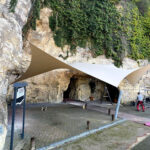Outdoor rock alcove with tan and white fabric canopies stretched overhead over a paved work area near a stone wall covered in vines.