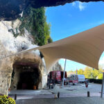 Rocky cave opening in a wall under a white fabric canopy; construction vans, bikes, and workers nearby.