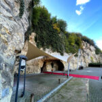 Rocky cliff with greenery and a cave-like entrance shaded by beige/white fabric sails; a red carpet leads to the cave.