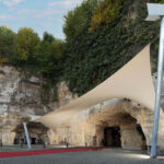 Rocky cliff face with ivy-covered wall and a cave entrance, shaded by a large beige fabric sail supported by metal poles, a red carpet leading to the cave, and a sign reading "Caverne" on the left.