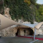 Outdoor rock wall with ivy-covered hillside and beige fabric shade sails anchored to metal poles over a red-carpeted walkway.
