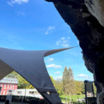 White tensile shade canopy extending from a rocky overhang over a paved courtyard, with a park, trees, and a red building in the background under a blue sky.