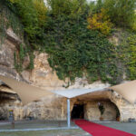 Rocky cliff covered in ivy and netting with a carved cave entrance; tan shade sails stretch over a red carpet path leading inside.