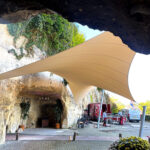 Outdoor area under a rocky overhang with a beige tensile shade, parked vans, and people at a setup site.