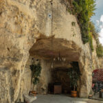 Open rock alcove converted into an indoor space with a chandelier hanging from the ceiling, potted plants, and a red van with bicycles nearby outside the entrance.