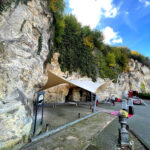 Rocky cliff face with ivy overhangs, set up for an outdoor event under a tan-and-white canopy, with ladders and equipment on a paved area.