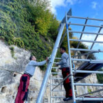 Two construction workers in grey shirts and red pants assemble a metal scaffolding frame beside a rocky wall outdoors.