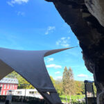 Shade sail anchored to a rocky overhang spans a courtyard, with a blue sky and trees in the background.
