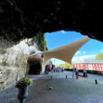 Outdoor plaza under a large white fabric shade attached to a rocky overhang, with a tunnel entrance and distant people and buildings.