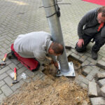 Two workers install a metal streetlamp pole in a paved area, securing the base with bolts while sand surrounds the foundation.