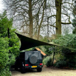 Dark SUV parked on a gravel driveway under a large black shade sail, with a spare tire cover on the rear and dense greenery around.