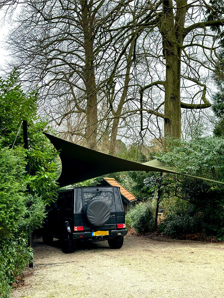 Dark SUV parked on a gravel driveway under a large black shade sail, with a spare tire cover on the rear and dense greenery around.