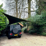 Dark SUV parked in a gravel driveway under a large black shade sail anchored between two posts in a garden setting.