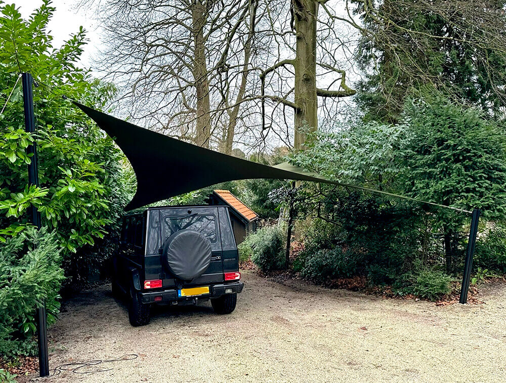 Dark SUV parked in a gravel driveway under a large black shade sail anchored between two posts in a garden setting.