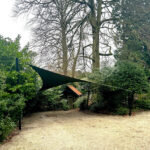 Green fabric shade sail stretched between two black posts over a gravel path in a garden with shrubs and trees in the background.