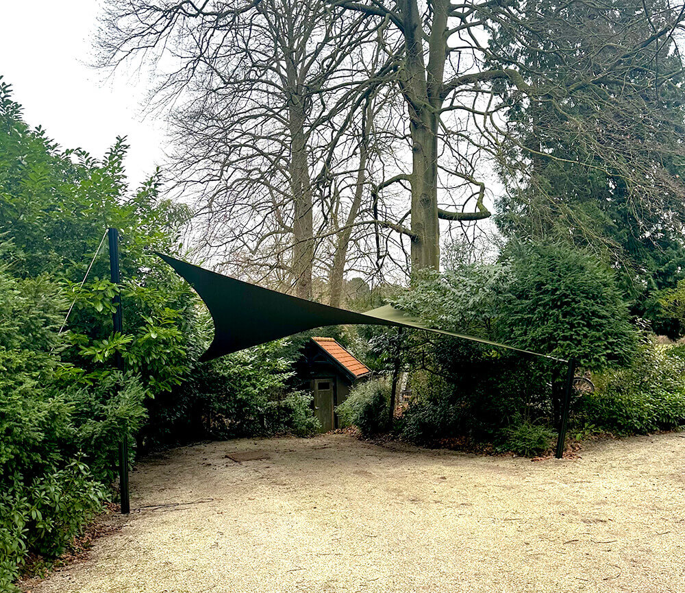 Green fabric shade sail stretched between two black posts over a gravel path in a garden with shrubs and trees in the background.