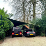 Two cars parked side by side under a triangular shade sail in a gravel driveway with green hedges and trees nearby.