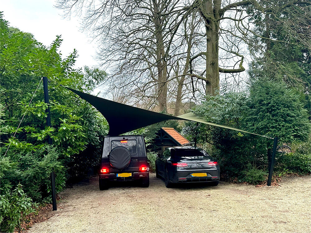 Two cars parked side by side under a triangular shade sail in a gravel driveway with green hedges and trees nearby.