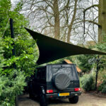 Black off-road SUV parked in a gravel driveway under a large triangular black shade sail, spare tire cover on the rear door, trees around it.