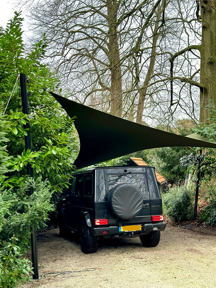 Black off-road SUV parked in a gravel driveway under a large triangular black shade sail, spare tire cover on the rear door, trees around it.