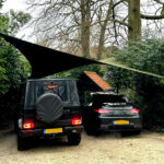 Two cars parked side by side under a large fabric shade sail stretched between tall posts in a gravel driveway, surrounded by green bushes and trees.