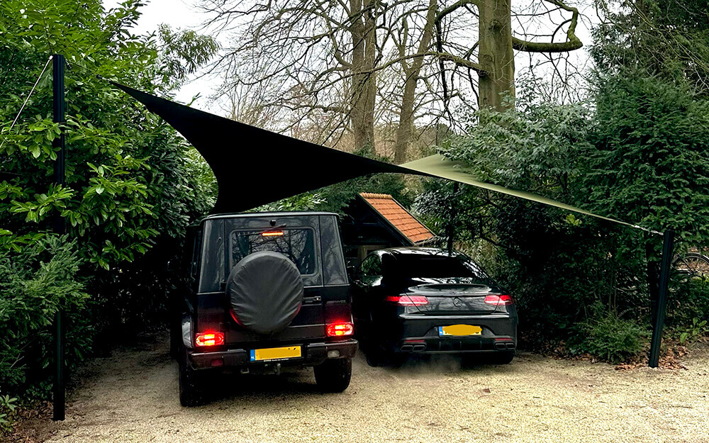 Two cars parked side by side under a large fabric shade sail stretched between tall posts in a gravel driveway, surrounded by green bushes and trees.