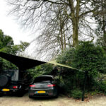 Two cars parked under a beige shade sail in a gravel lot, with green bushes nearby and a tall bare tree overhead.