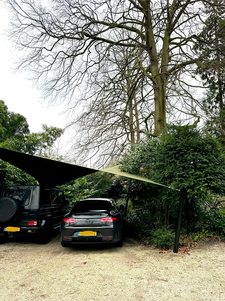 Two cars parked under a beige shade sail in a gravel lot, with green bushes nearby and a tall bare tree overhead.