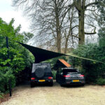 Shade sail covers two parked cars in a gravel driveway, supported by posts among greenery, providing shade in a garden setup.