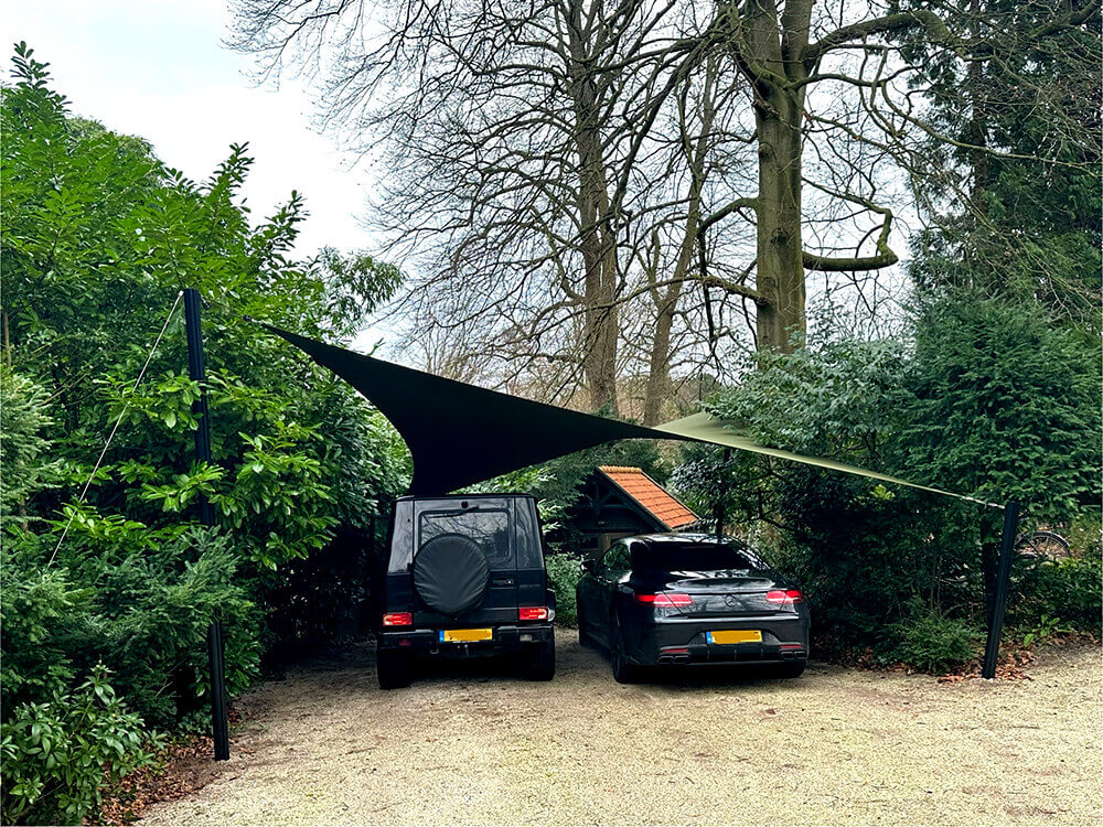 Shade sail covers two parked cars in a gravel driveway, supported by posts among greenery, providing shade in a garden setup.