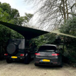 Two cars parked on a gravel driveway under a large triangular shade sail, surrounded by greenery and trees.