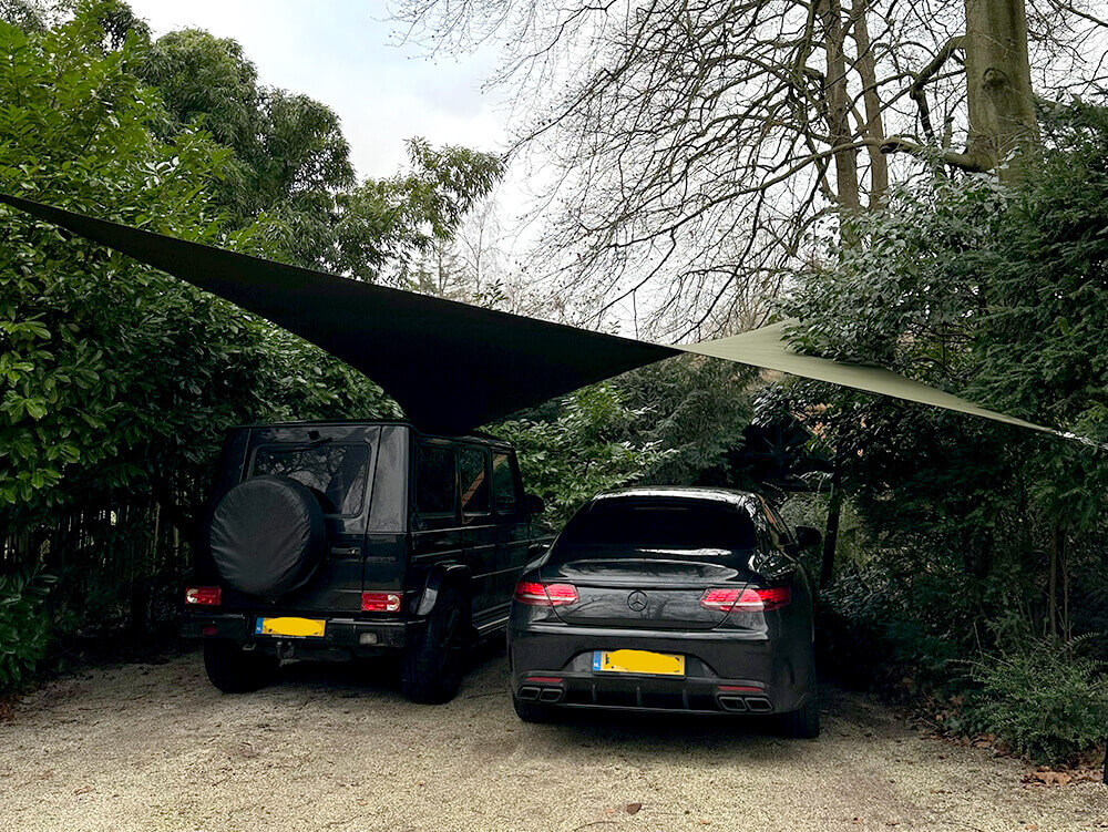 Two cars parked on a gravel driveway under a large triangular shade sail, surrounded by greenery and trees.