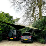 Two cars parked side by side under a triangular shade sail in a gravel driveway, surrounded by hedges and trees.