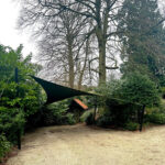 Shade sail stretched between two posts over a gravel path in a garden, with tall bare trees and dense shrubbery behind it.