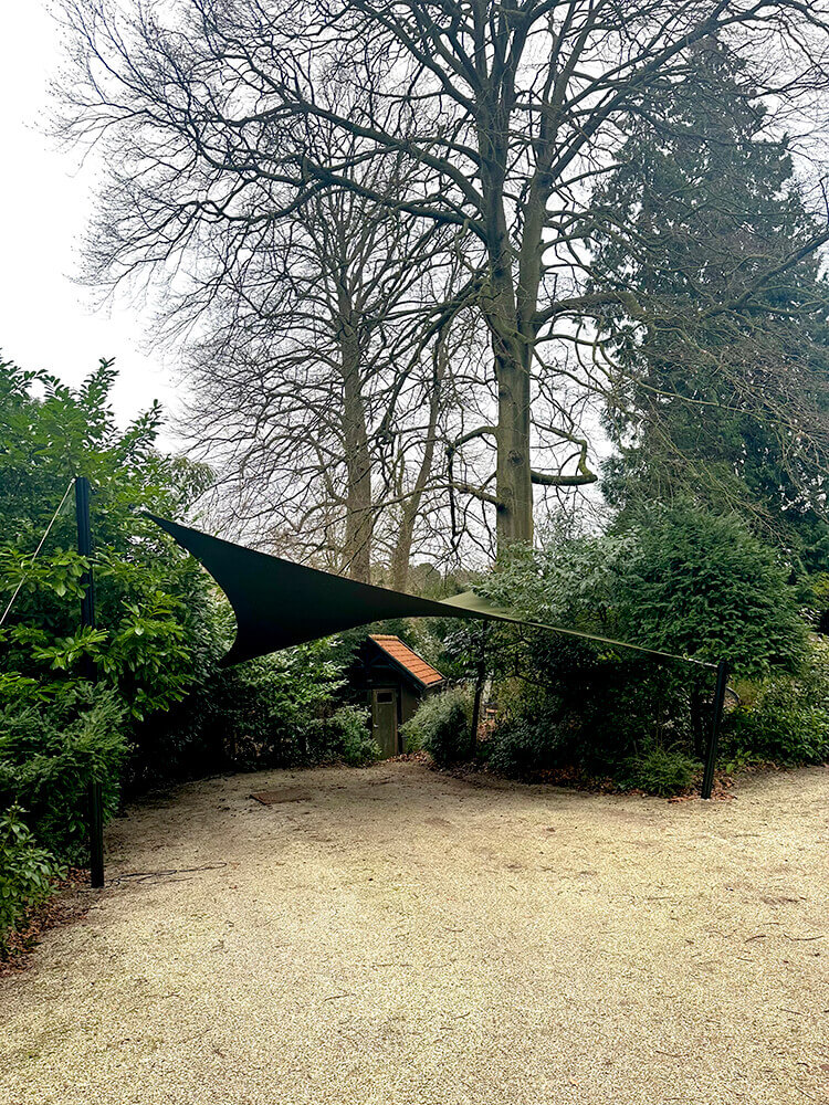 Shade sail stretched between two posts over a gravel path in a garden, with tall bare trees and dense shrubbery behind it.
