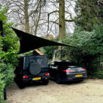 Two dark cars parked on a gravel driveway beneath a black triangular shade sail, surrounded by trees and green hedges.