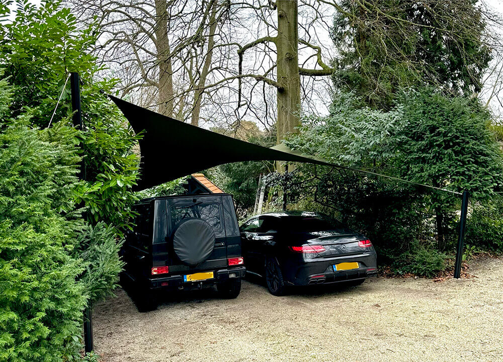 Two dark cars parked on a gravel driveway beneath a black triangular shade sail, surrounded by trees and green hedges.