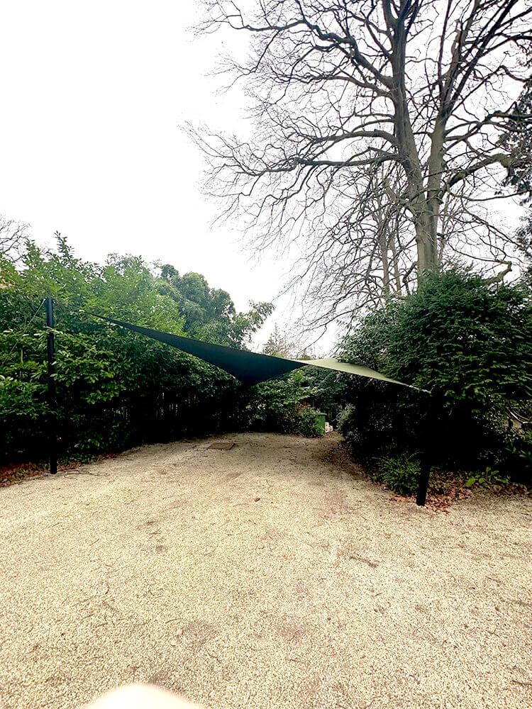 Gravel garden path flanked by hedges and a green triangular shade sail overhead.