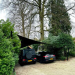 Two dark cars parked under a triangular black shade sail beside hedges and tall trees in a gravel lot.