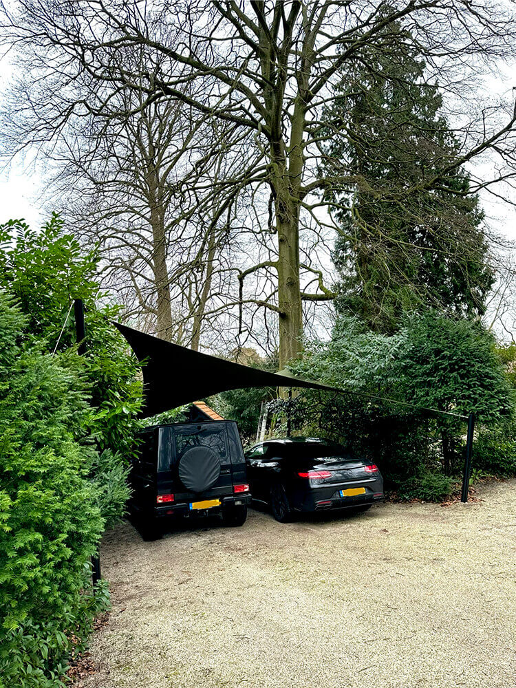 Two dark cars parked under a triangular black shade sail beside hedges and tall trees in a gravel lot.