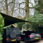 Two cars parked on a gravel driveway under a large black shade sail, surrounded by trees and greenery.