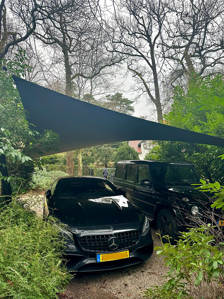 Two dark-colored cars parked under a large blue triangular shade sail in a lush garden, with bare trees in the background.