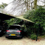 A black sedan parked under a tan fabric shade sail between trees and bushes in a gravel lot