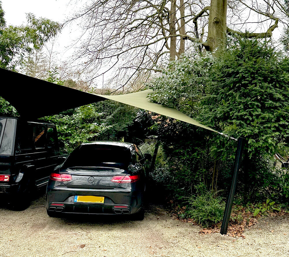 A black sedan parked under a tan fabric shade sail between trees and bushes in a gravel lot