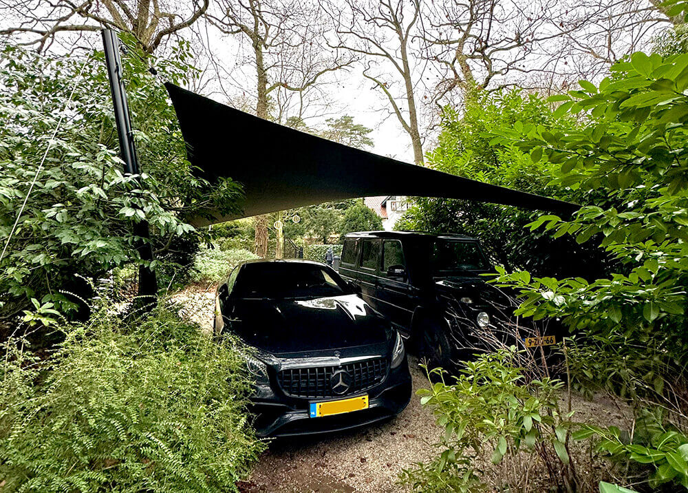 Two black cars parked under a large shade sail in a overgrown garden area, with dense shrubs and trees surrounding them