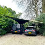 Two cars parked side by side under a fabric shade sail in a gravel driveway, surrounded by dense green hedges and trees.