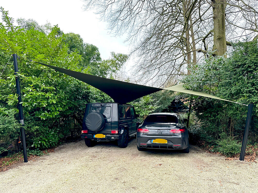 Two cars parked side by side under a fabric shade sail in a gravel driveway, surrounded by dense green hedges and trees.